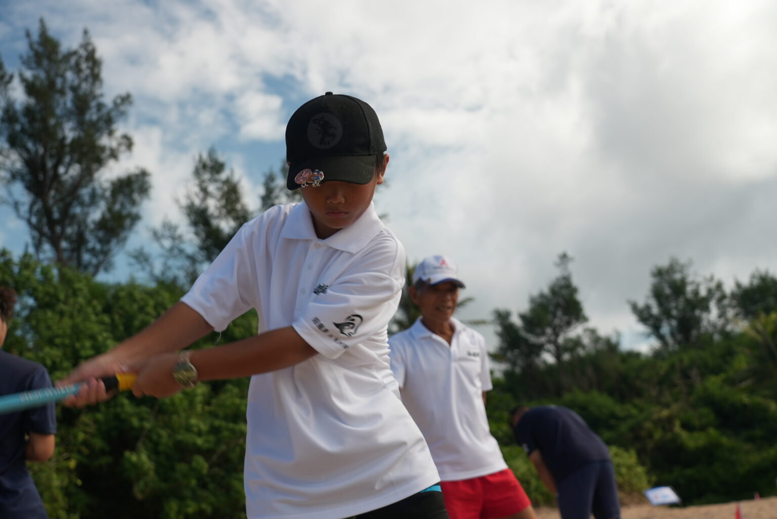 青少年選手在沙灘球道上揮桿擊球，身後有一位年長選手與其他參賽者，背景為海岸林
