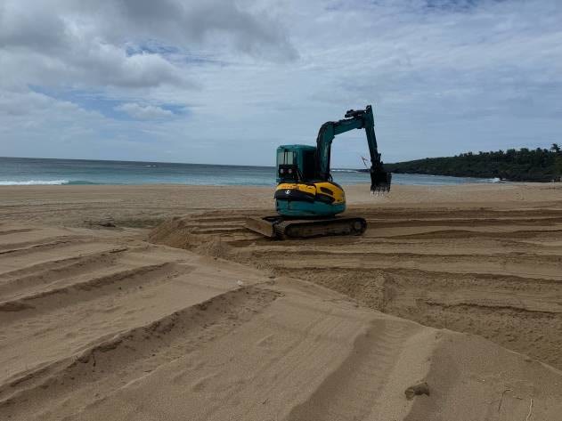 重型機具停在白沙灣沙灘上，周圍是整平後的沙地痕跡，遠方為海面與海岸林